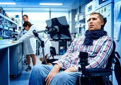A man in a wheelchair sits in a lab, with two scientists in the background